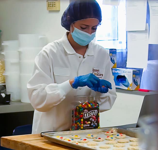 Baker hand-decorating freshly baked cookies with chocolate toppings in Jimmy&rsquo;s Bakery production kitchen