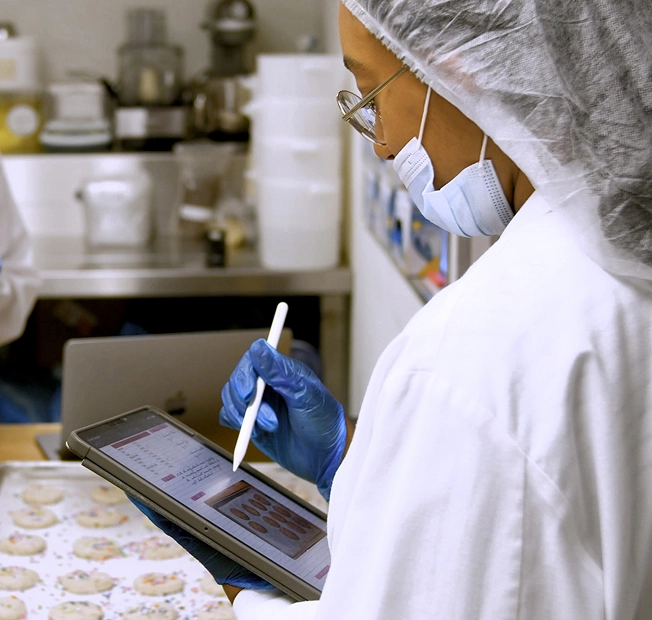 Food safety technician in hairnet and mask recording quality checks on a tablet in a cookie facility.